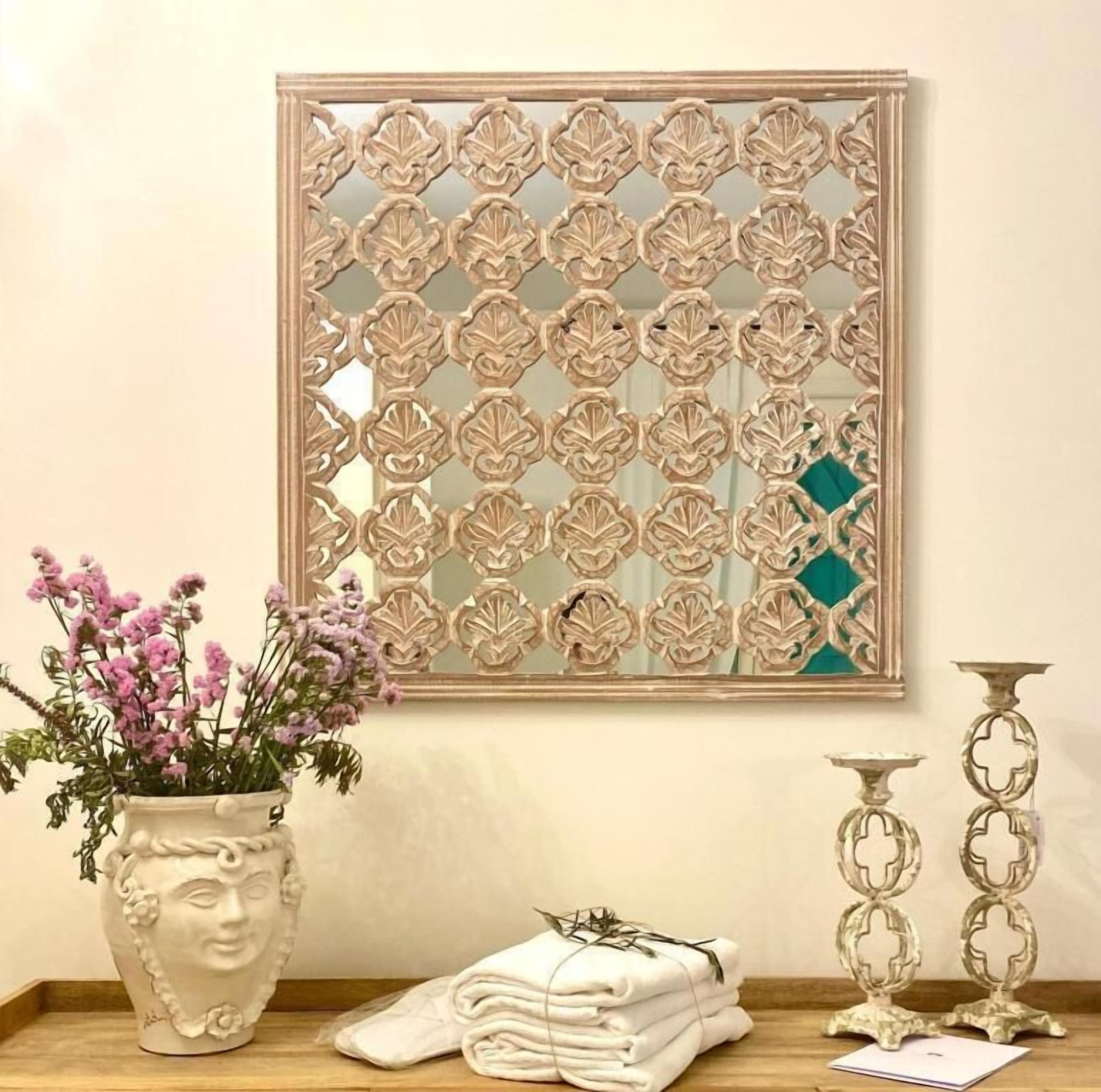 Espelho decorativo com padrão floral, um vaso com flores, toalhas dobradas e castiçais sobre uma superfície de madeira.