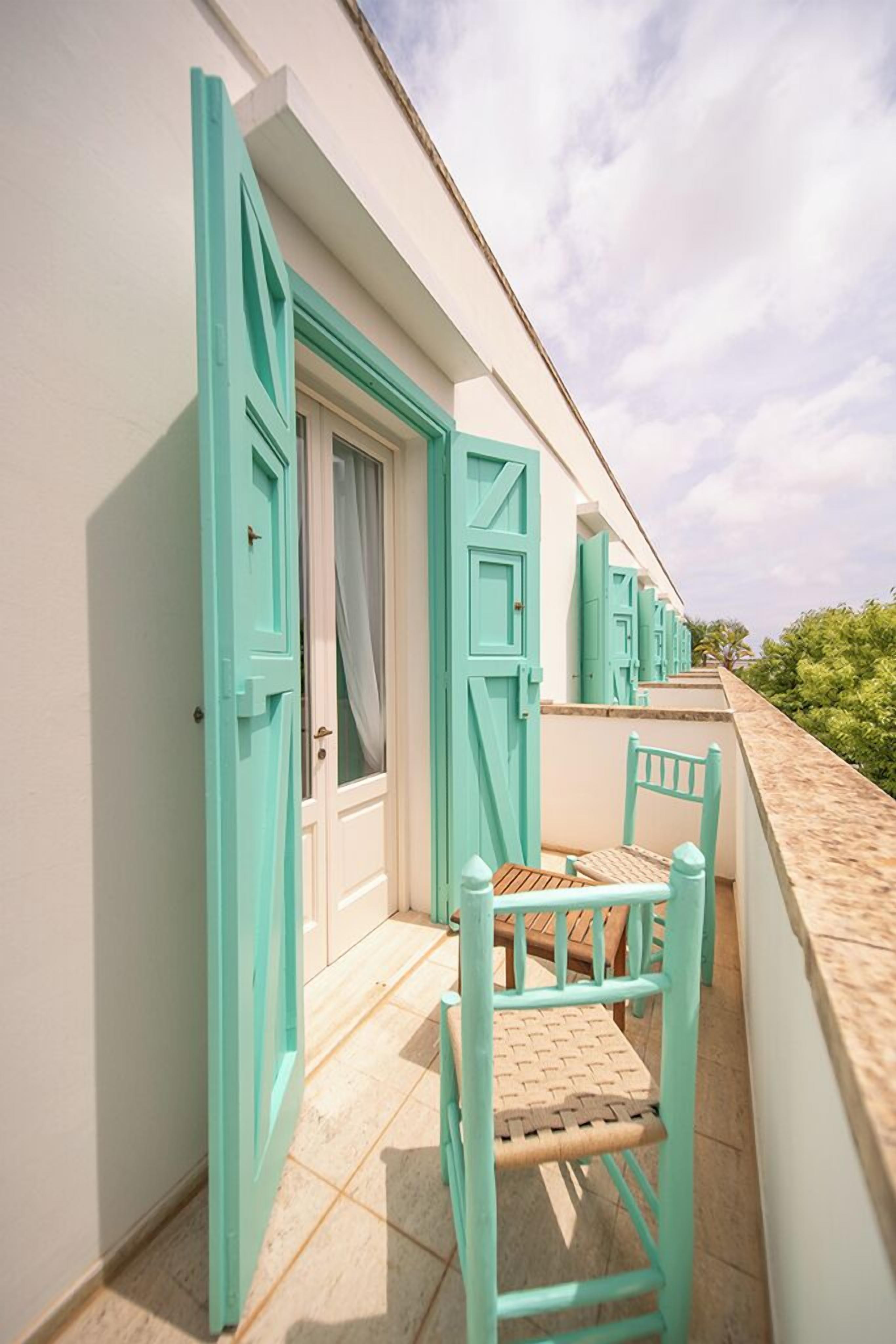 Een balkon met blauwgroene deuren en stoelen, een betegelde vloer, en een heldere lucht.