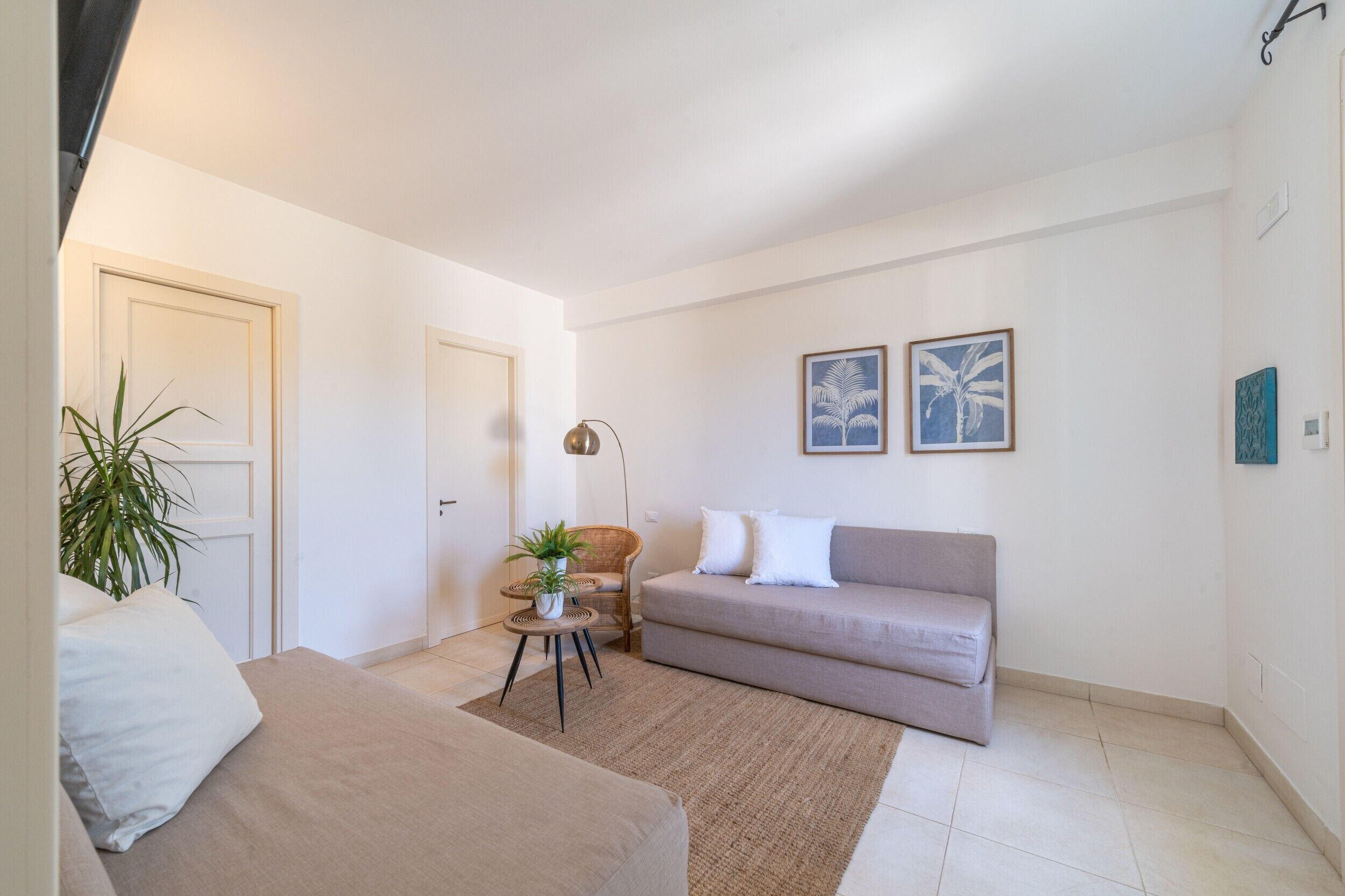A living room with a beige sofa, a small round table with a plant, and a floor lamp. There are two framed pictures on the wall and a door with a plant beside it.