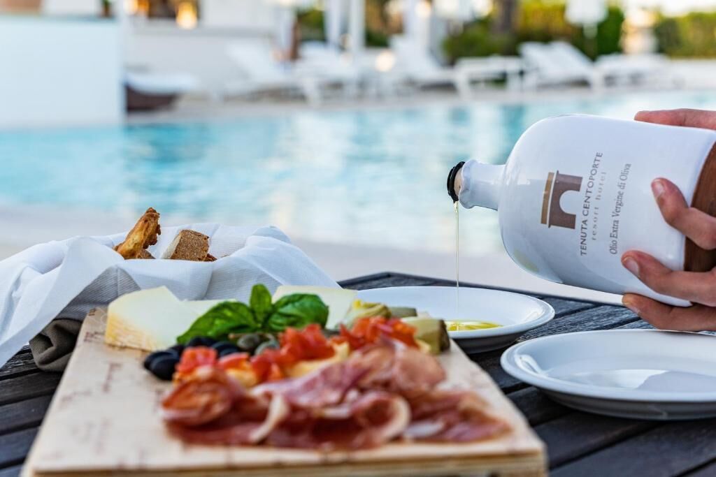 A person pouring olive oil over a charcuterie board with various meats and cheeses by a poolside.