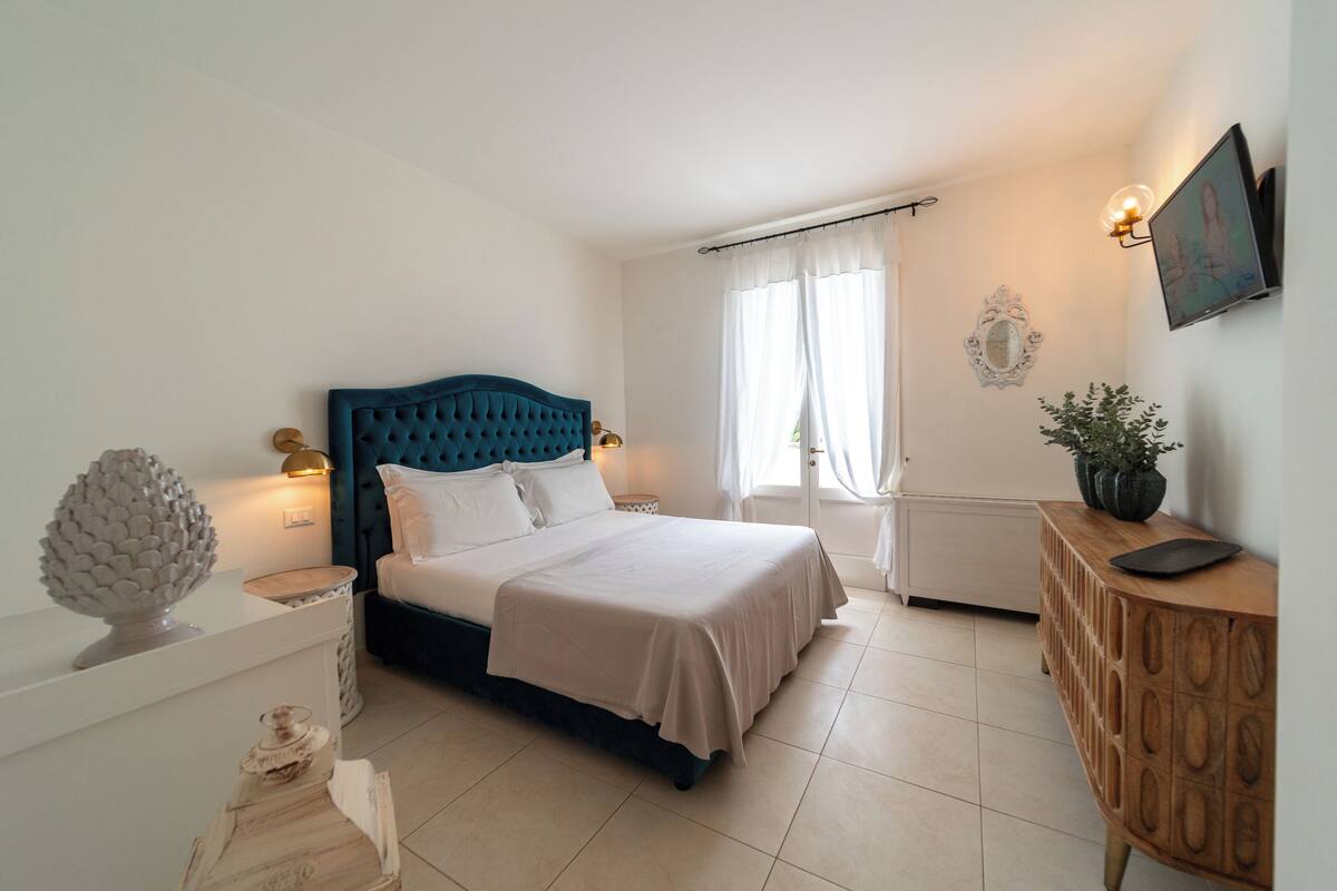A bedroom with a bed, bedside tables, a dresser, a window with curtains, and a wall-mounted TV.
