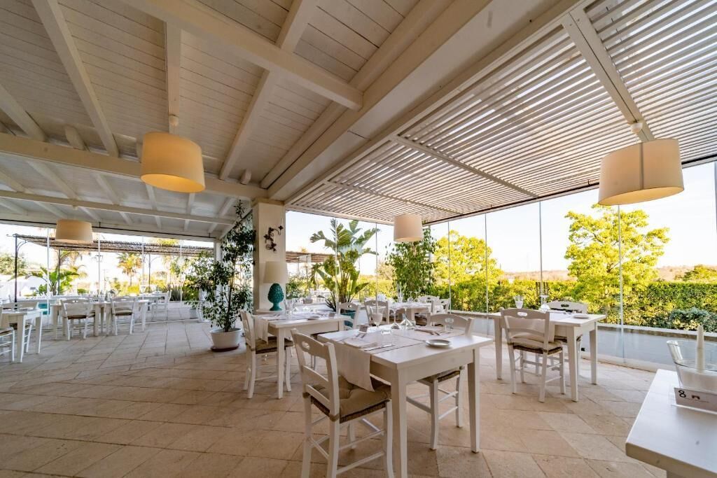 Un área de comedor al aire libre con mesas y sillas blancas, plantas en macetas y lámparas colgantes.