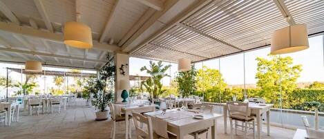 Un espace extérieur aménagé pour les repas, avec des tables et des chaises blanches, des plantes en pot et des lampes suspendues.
