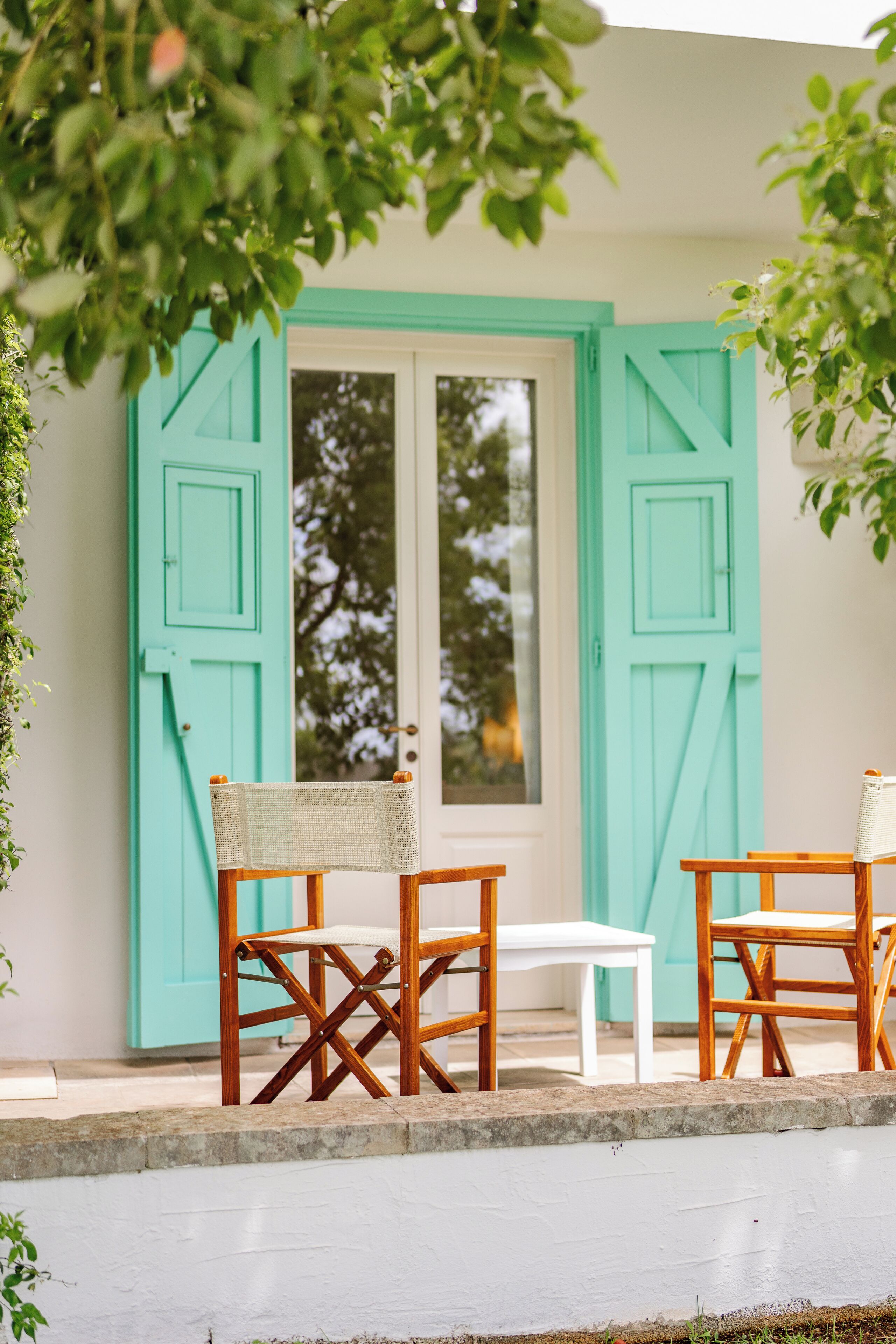 Una puerta turquesa con un ventanal blanco, dos sillas de madera con asientos de mimbre y una pequeña mesa blanca.