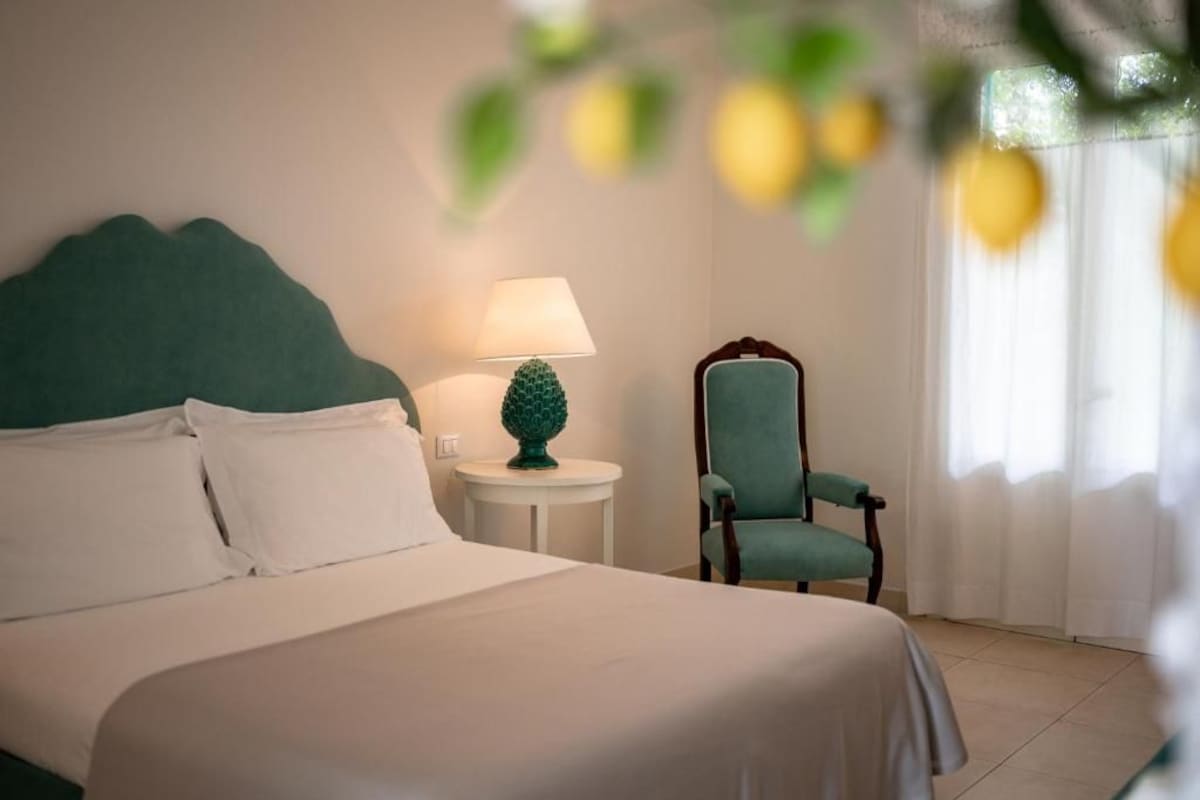 Una cama bien hecha con sábanas blancas, un cabecero tapizado en verde, una mesita redonda con una lámpara verde y una silla color aguamarina.