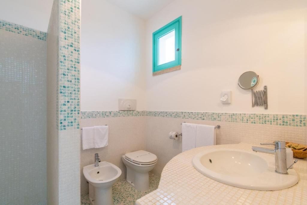 A bathroom with a white sink, a toilet, and a bidet. There is a small window with a turquoise frame and a mirror on the wall.