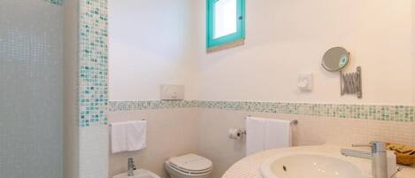 A bathroom with a white sink, a toilet, and a bidet. There is a small window with a turquoise frame and a mirror on the wall.