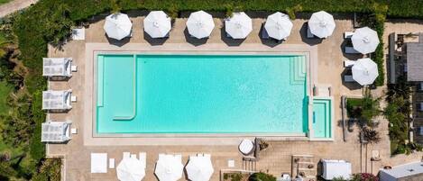 Vista aérea de una piscina rectangular rodeada de sombrillas blancas y sillas de descanso, con un jardín ajardinado y un paseo pavimentado.