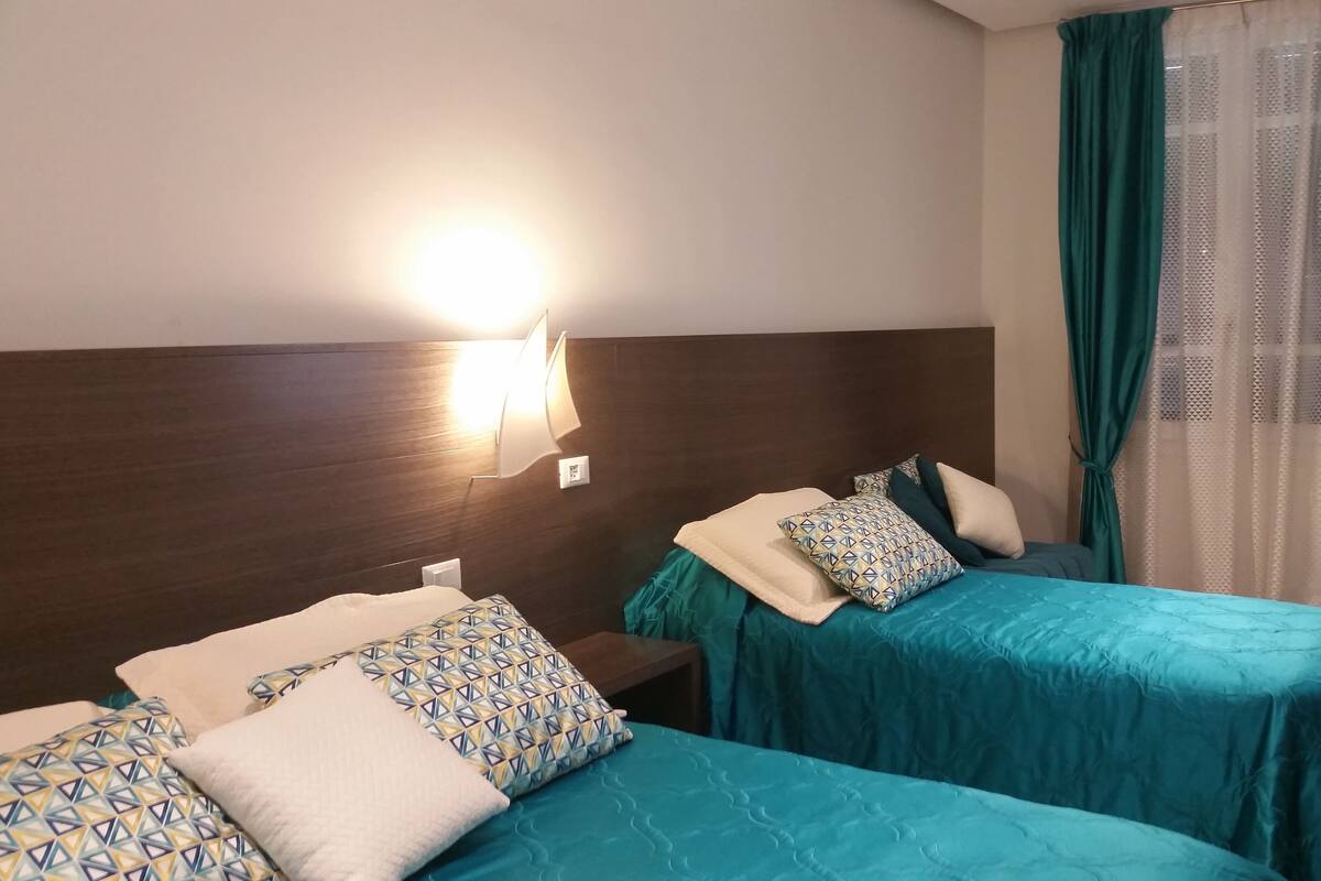 Hotelkamer met twee bedden, elk voorzien van kussens met een patroon, een hoofdbord, een wandlamp en een raam met gordijnen.