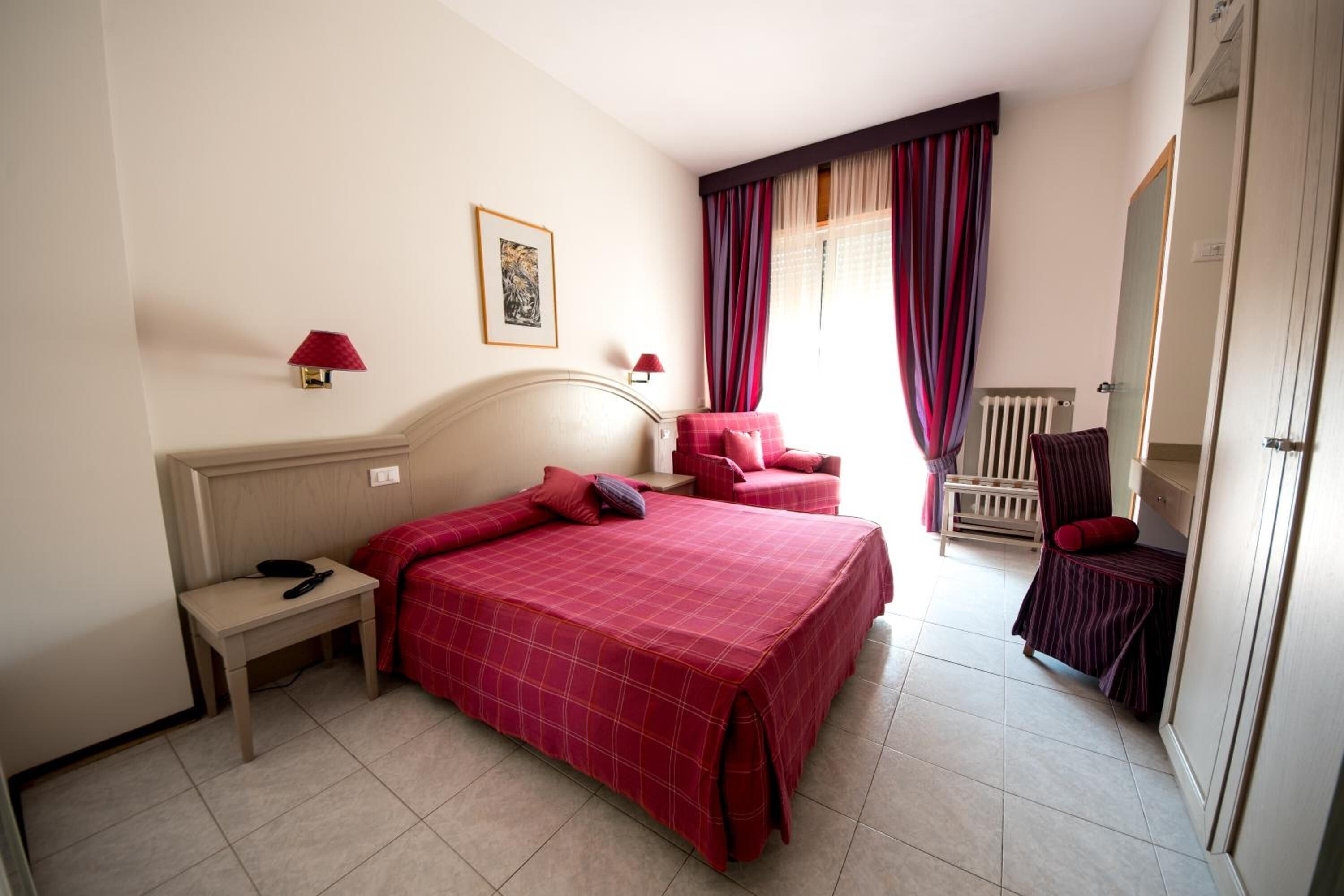 Une chambre d’hôtel avec un lit, une table de chevet, une lampe, une chaise et une fenêtre avec des rideaux.