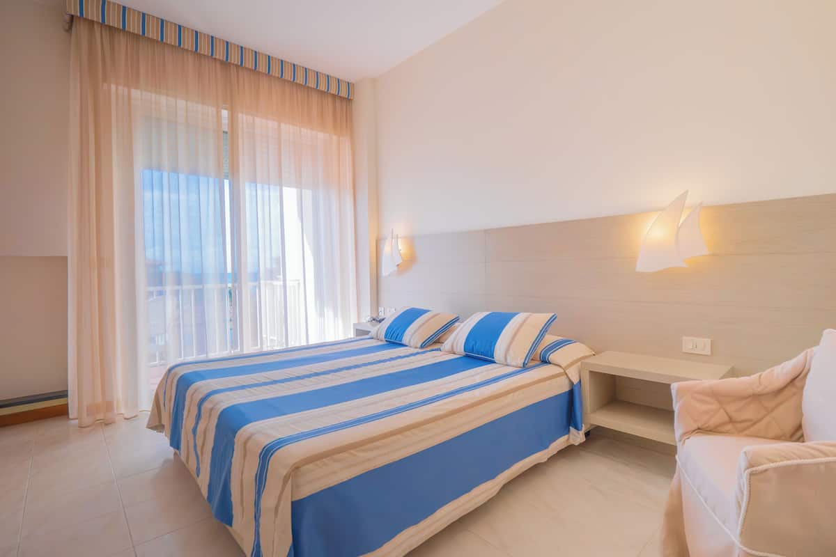 Een hotelkamer met een bed, nachtkastje, stoel en raam met dunne gordijnen.