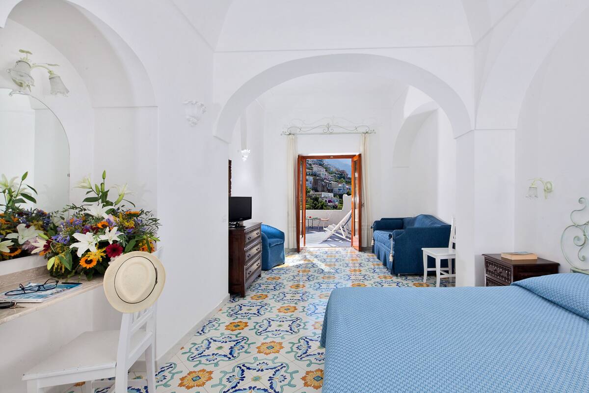 A hotel room with a bed, a desk with a hat and glasses, a chair, a dresser, and a view of the outdoors.