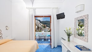 Una habitación de hotel con una cama, un escritorio con un libro, un televisor, un espejo y un balcón con vista a un pueblo.