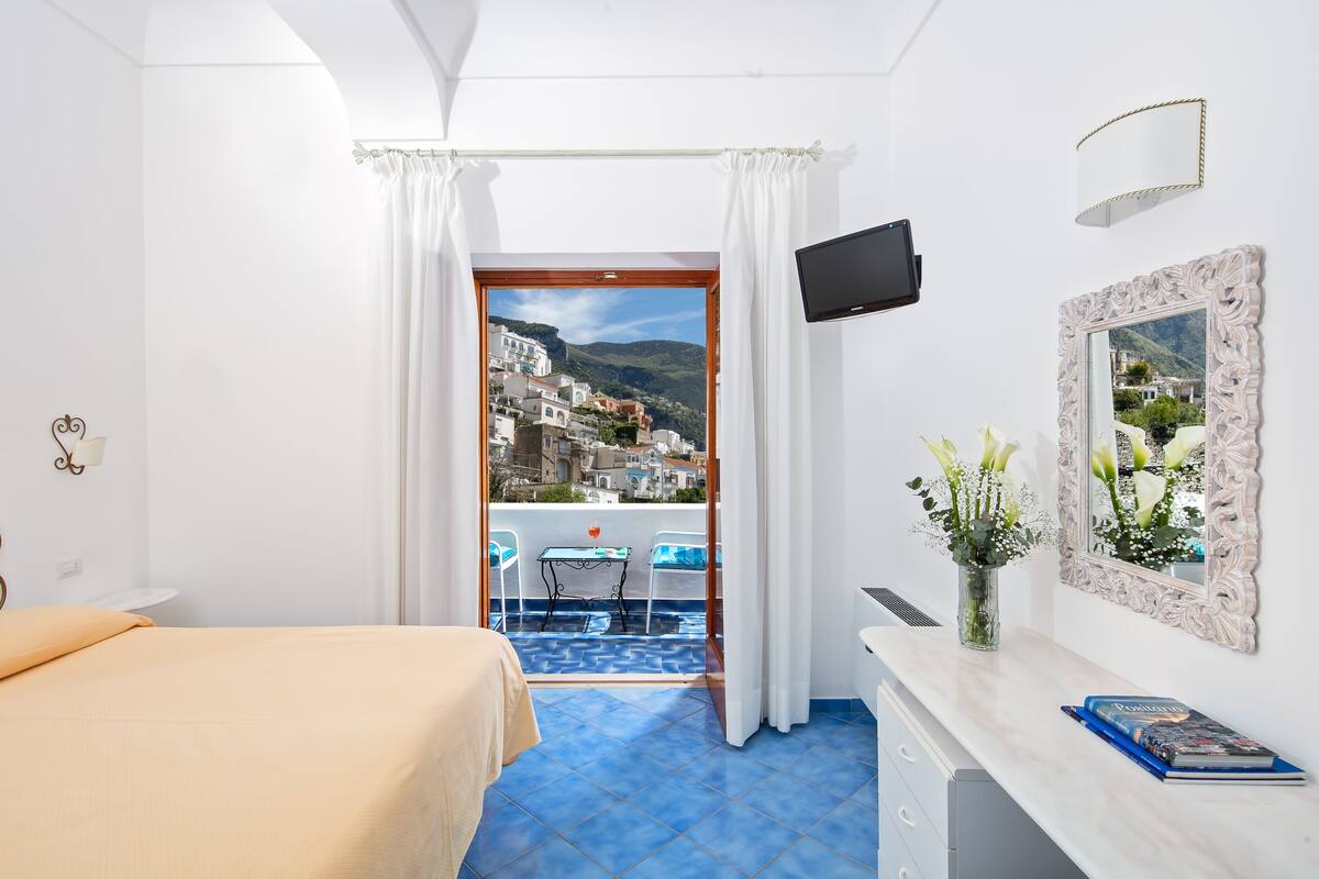 A hotel room with a bed, a desk with a book, a TV, a mirror, and a balcony with a view of a town.