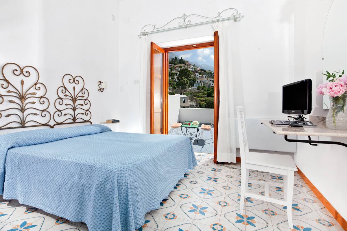 A bedroom with a bed, a desk with a computer, and a view of a hillside.