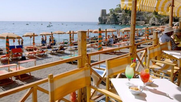 Un restaurante frente a la playa con terraza al aire libre, con vistas al mar y acantilados rocosos.