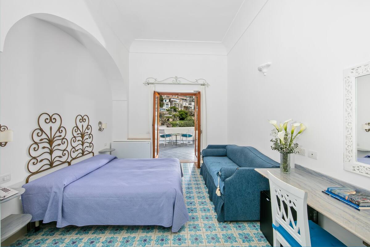 A hotel room with a bed, a sofa, a desk, and a view through an open door.