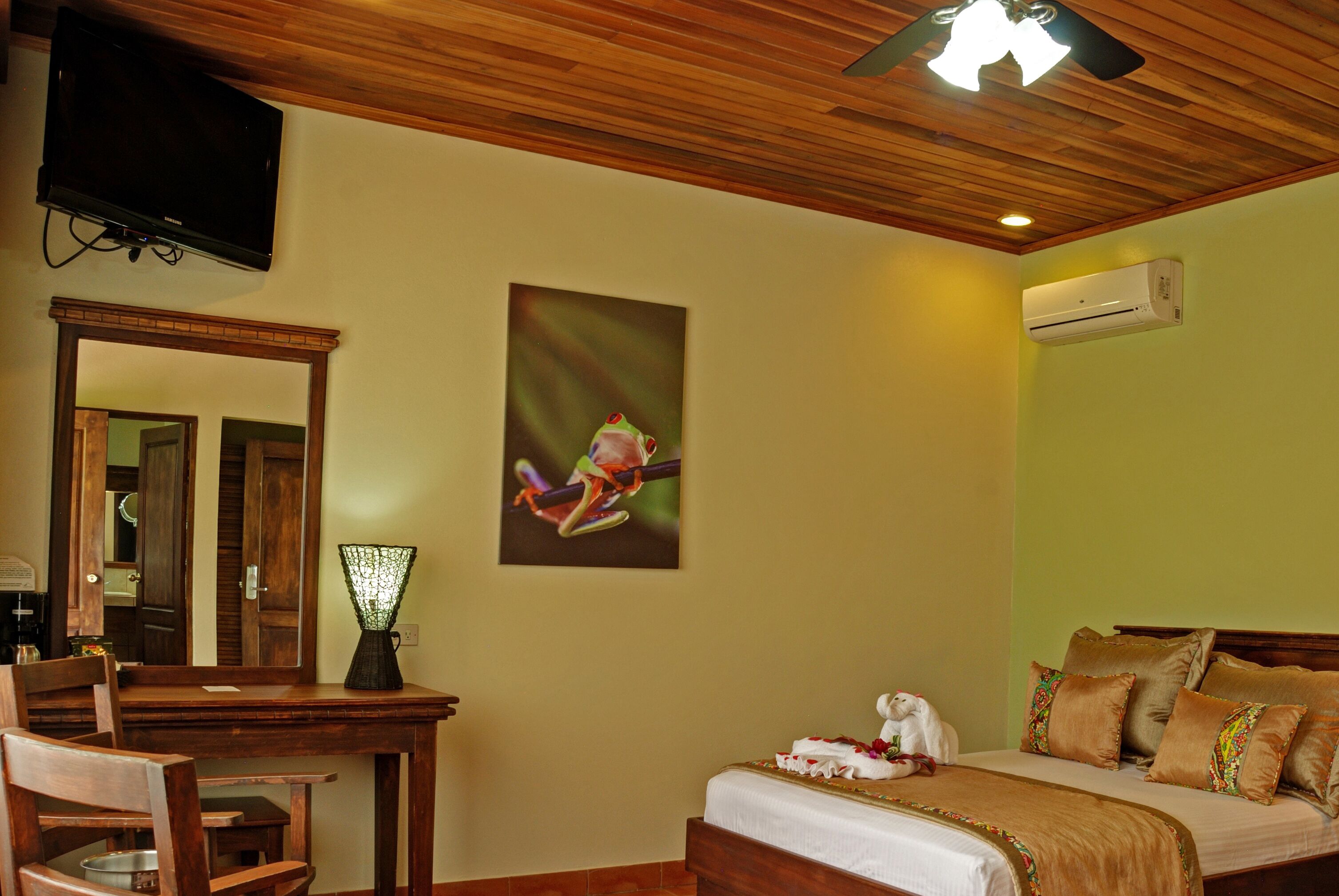 Habitación de hotel con techo de madera, televisor de pantalla plana en la pared, cama con ropa de cama marrón, escritorio con silla y espejo.