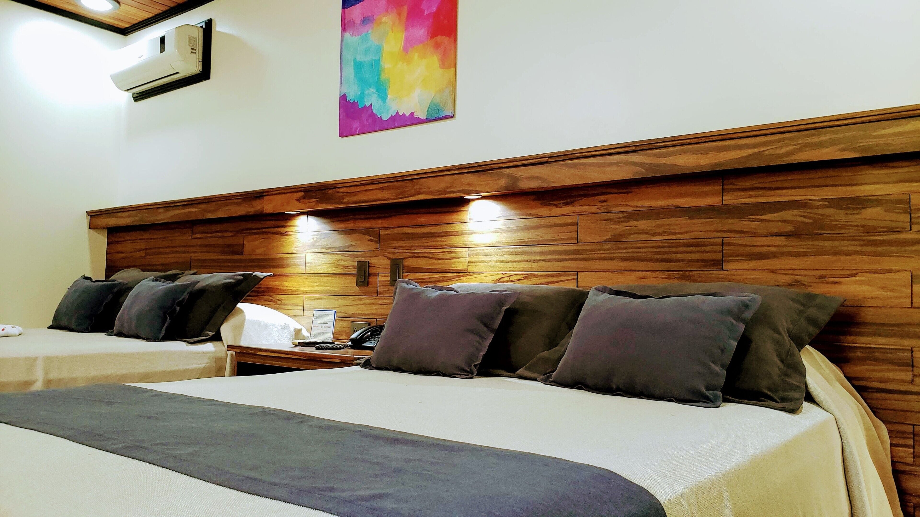 A hotel room with two beds, wooden headboards, a nightstand with a phone and a lamp, and a colorful abstract painting on the wall.