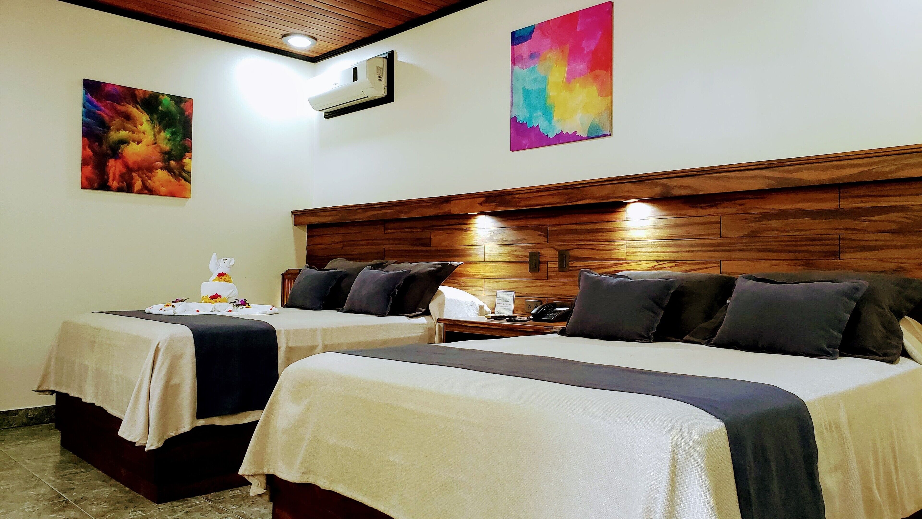 A hotel room with two beds, wooden headboards, a small table with a phone and a menu, and artwork on the walls.