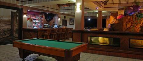 A pool table in a room with a bar area, wooden chairs, and a decorative rock feature.