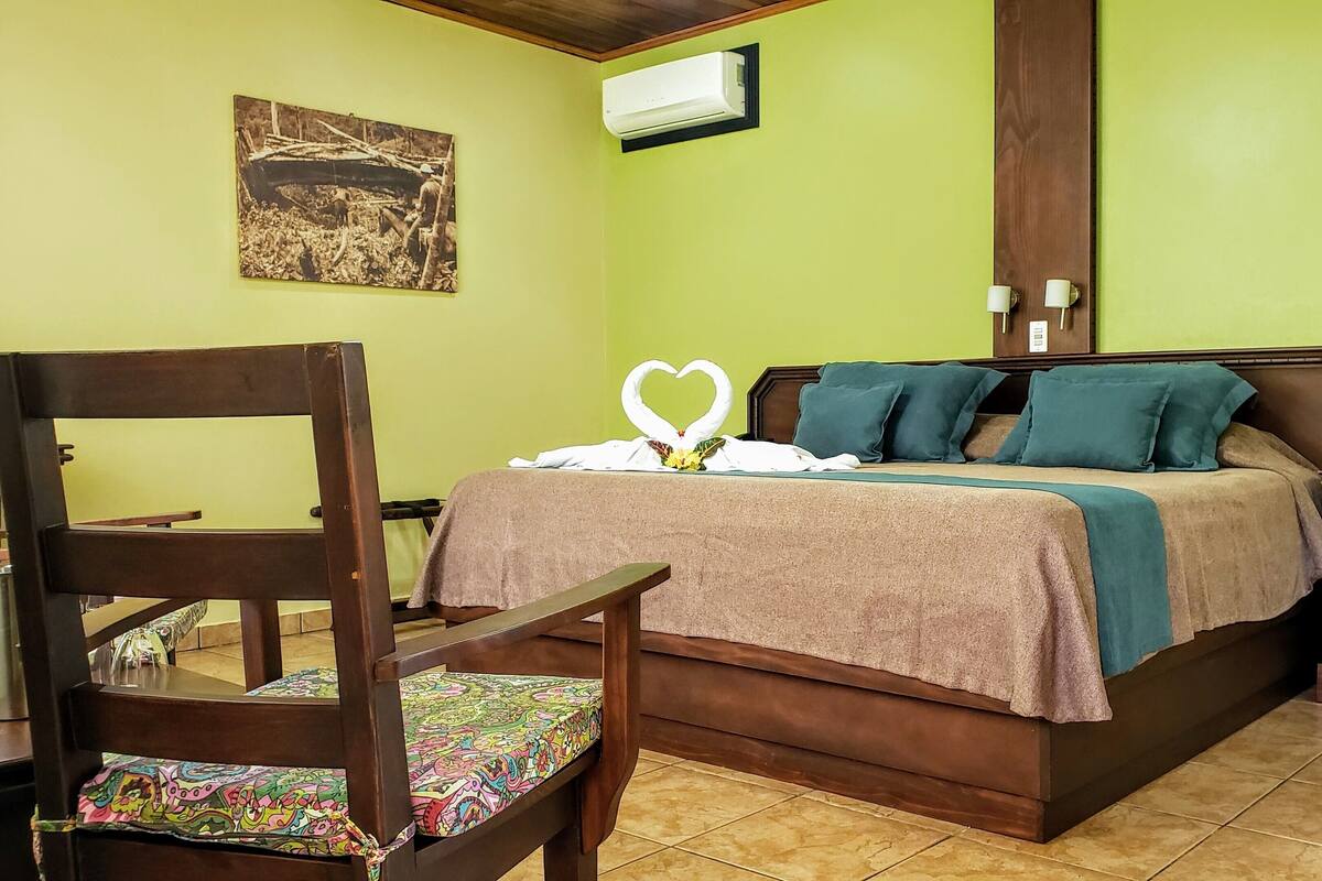 Une chambre d’hôtel avec un lit, une chaise en bois, une table de chevet et un climatiseur fixé au mur.