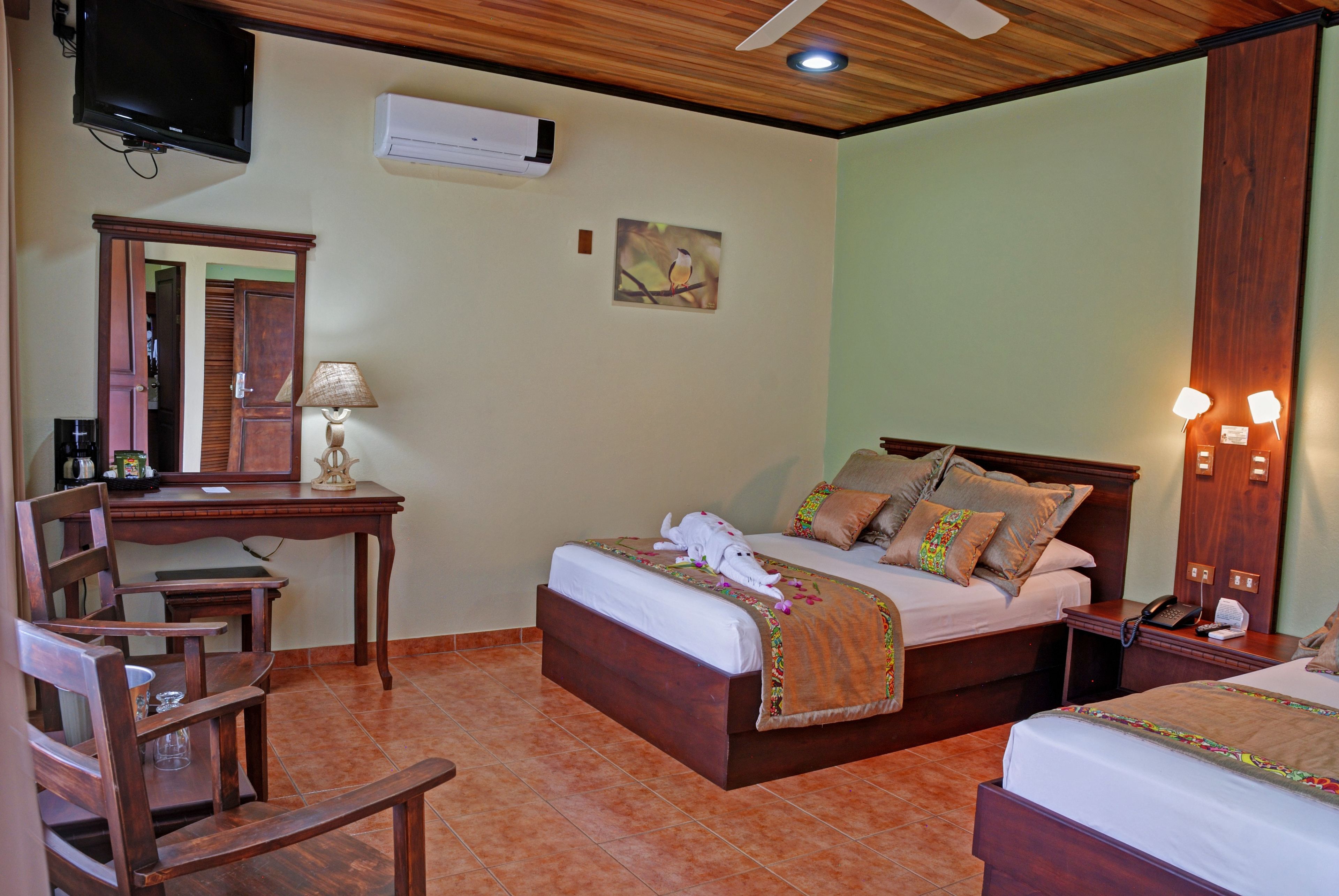 A hotel room with a wooden bed, a desk with a lamp, a television, and a wall-mounted air conditioner.