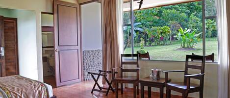 A bedroom with a bed, a window with a view of a garden, a wooden ceiling, and a door.