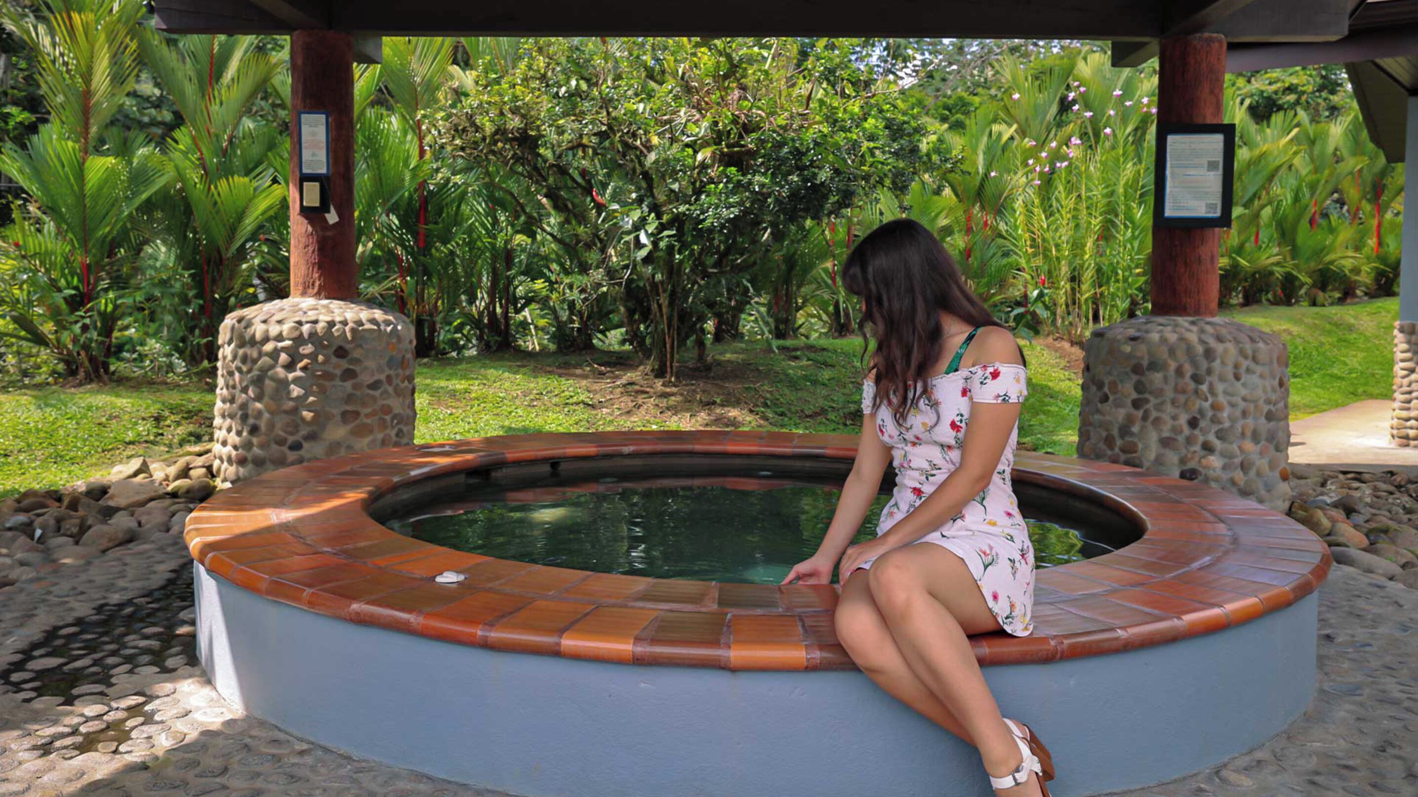 A woman sitting by a circular hot tub with stone pillars, surrounded by lush greenery.