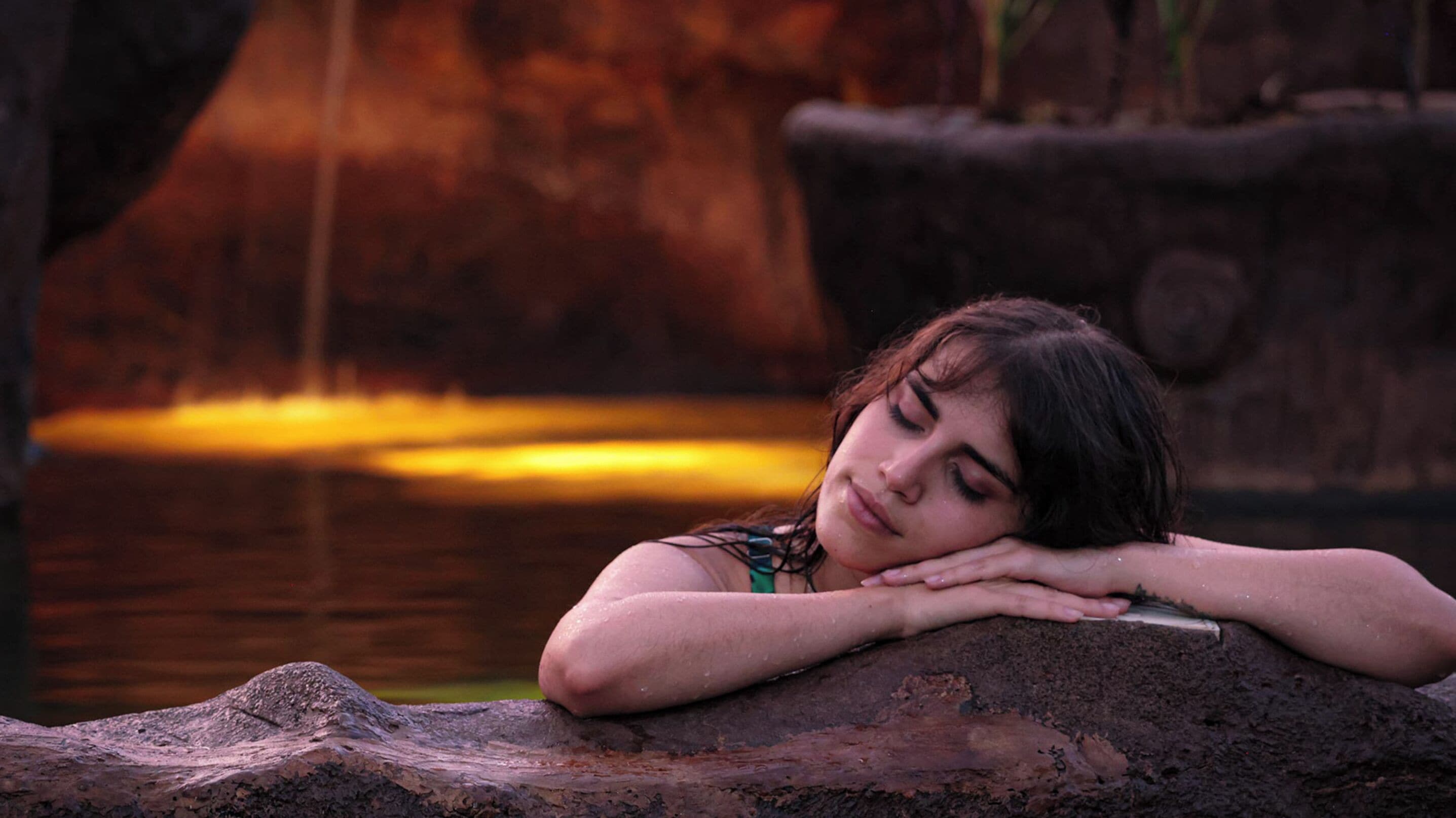 A person resting on a rock by a body of water with a waterfall in the background.
