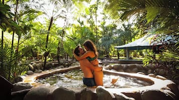 Um casal se abraçando em um cenário natural com um espelho d'água e vegetação exuberante.