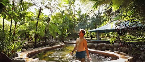 A man in a natural setting with a water feature surrounded by rocks and lush greenery.