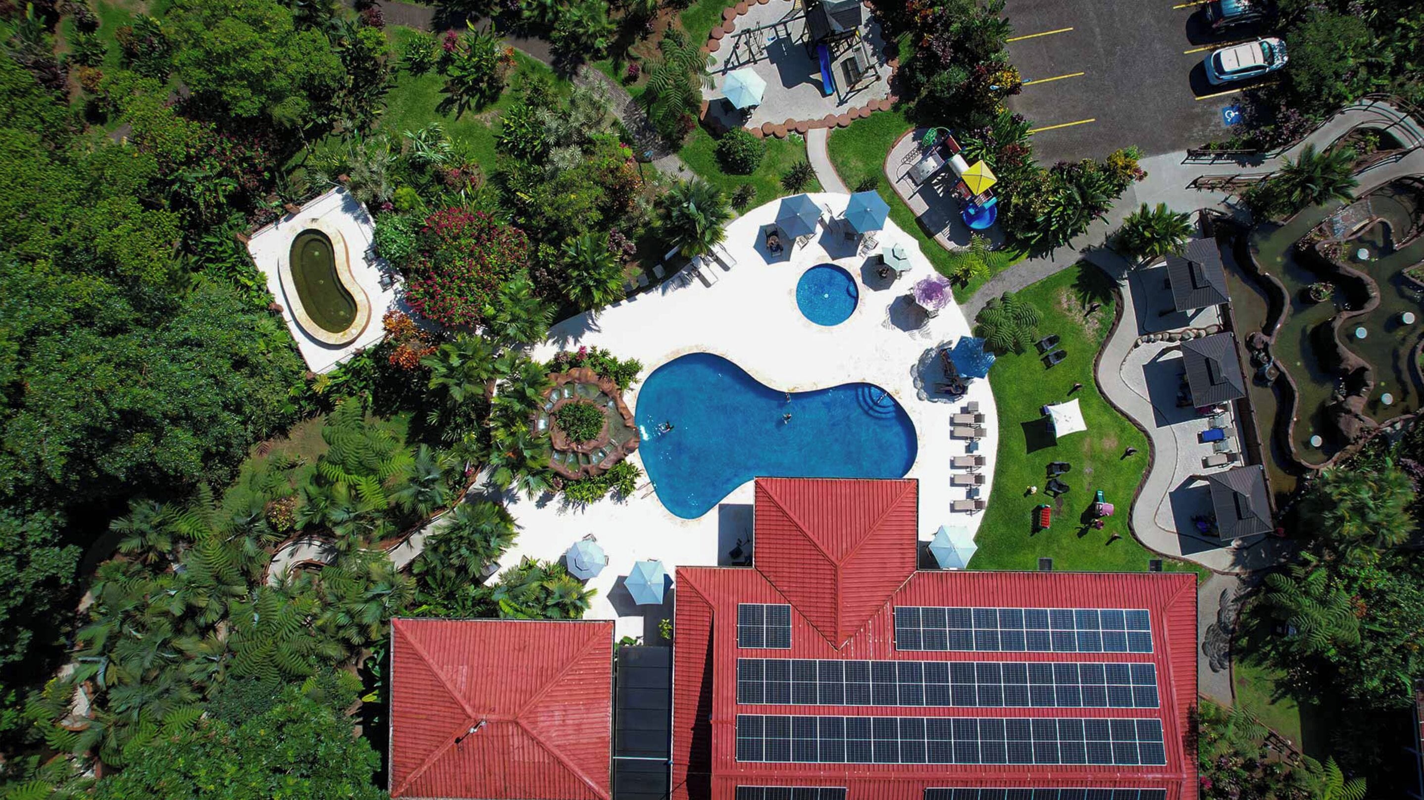 Vista aérea de um resort com uma piscina grande, espreguiçadeiras e um edifício com telhado vermelho e painéis solares.