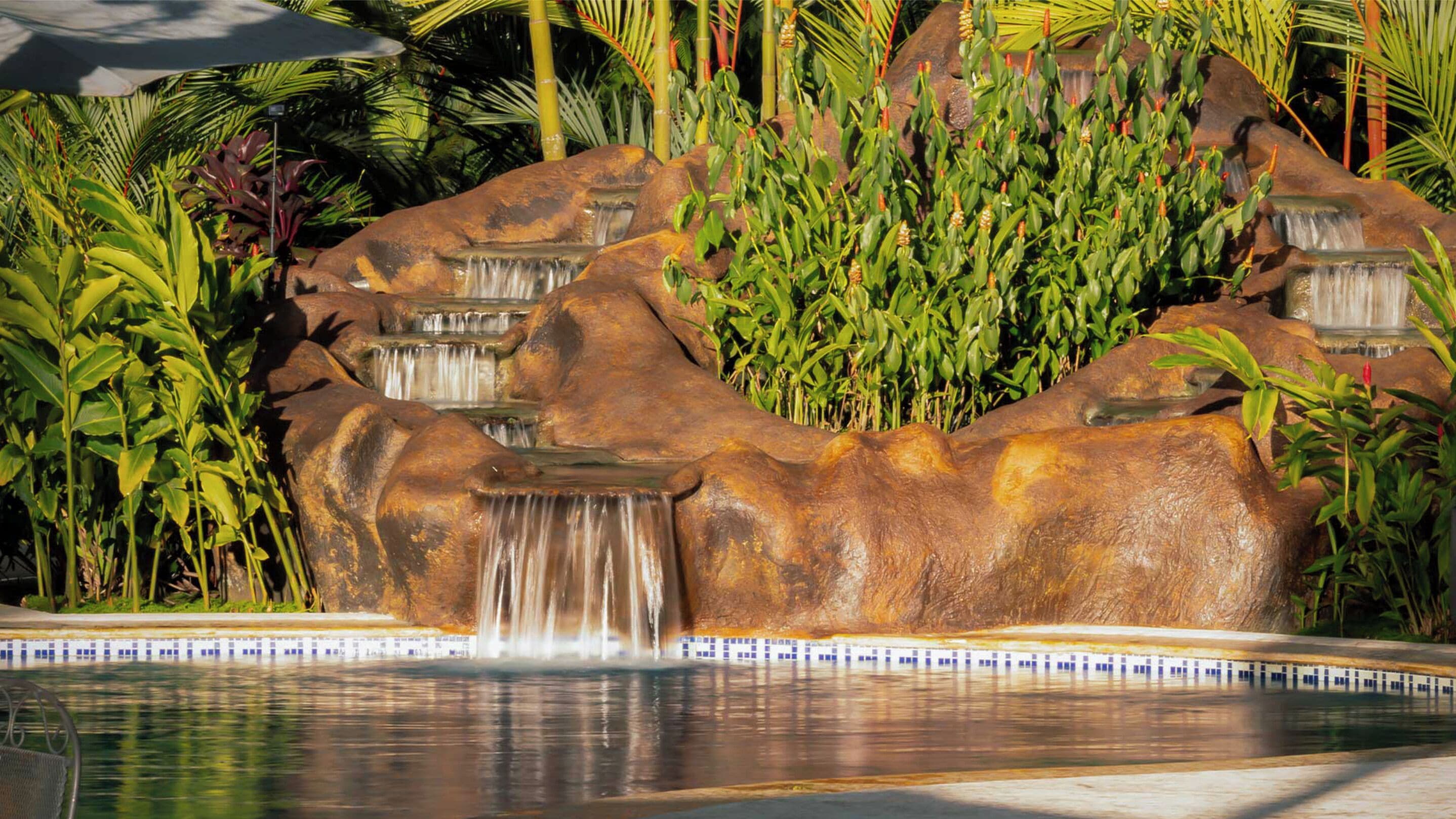 A rock waterfall feature with a pool in front, surrounded by tropical plants.