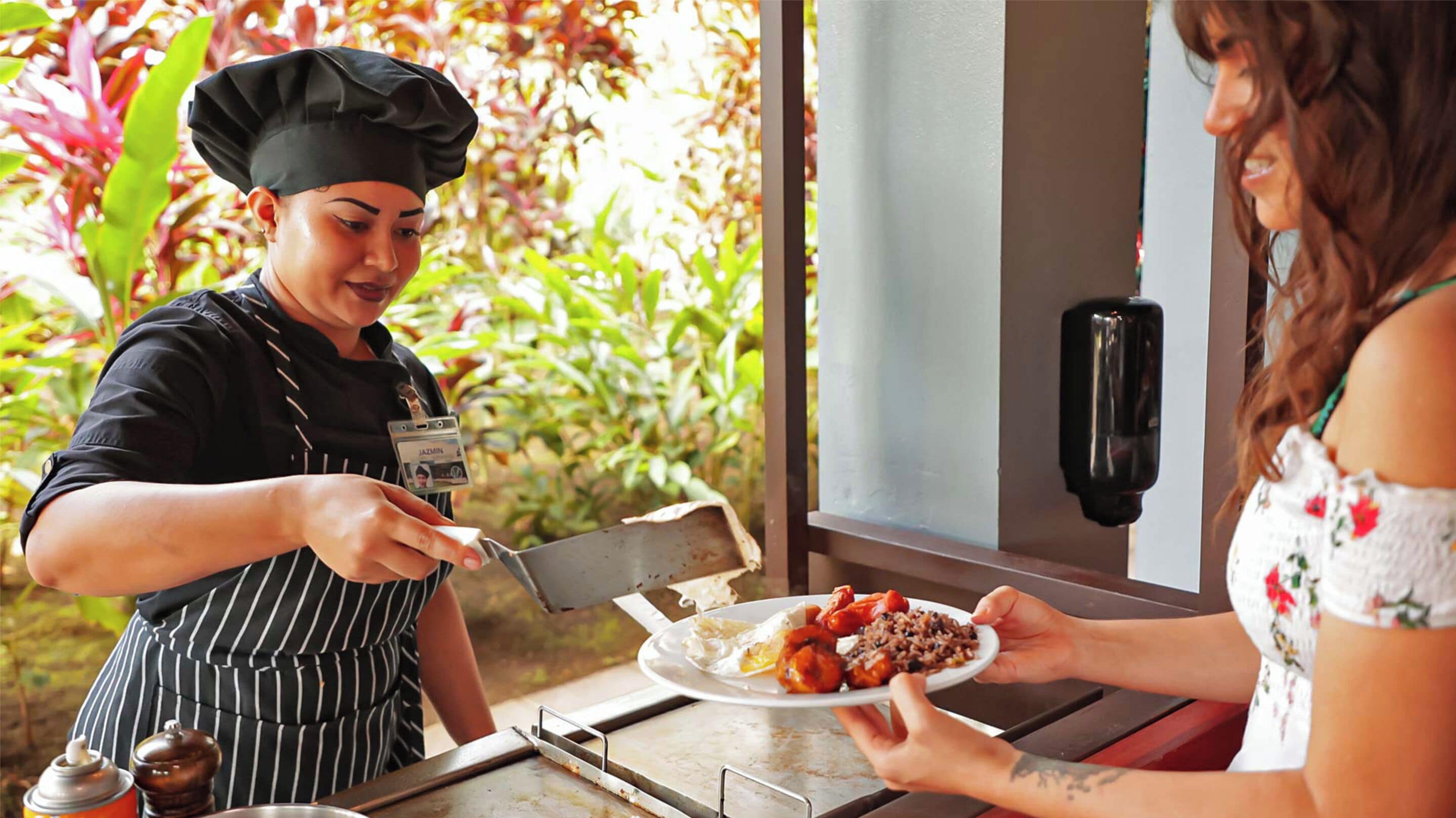 A person in a chef's uniform serving food to another person at an outdoor table.