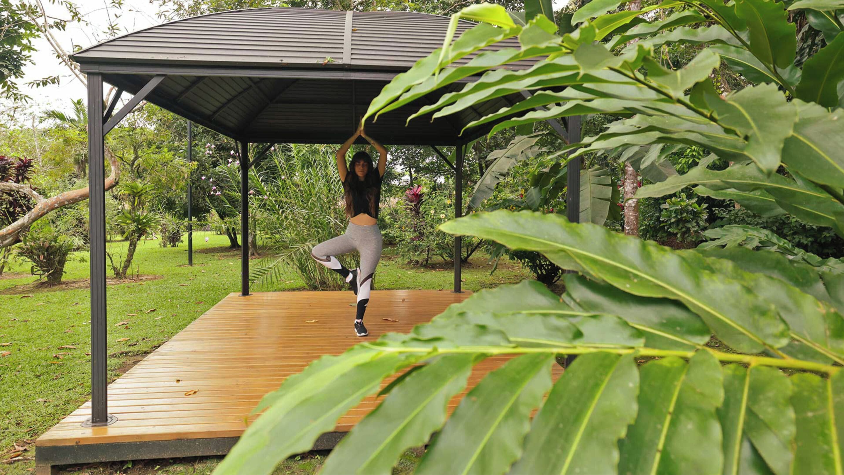 A person performing an exercise under a wooden deck with a metal roof, surrounded by lush greenery.