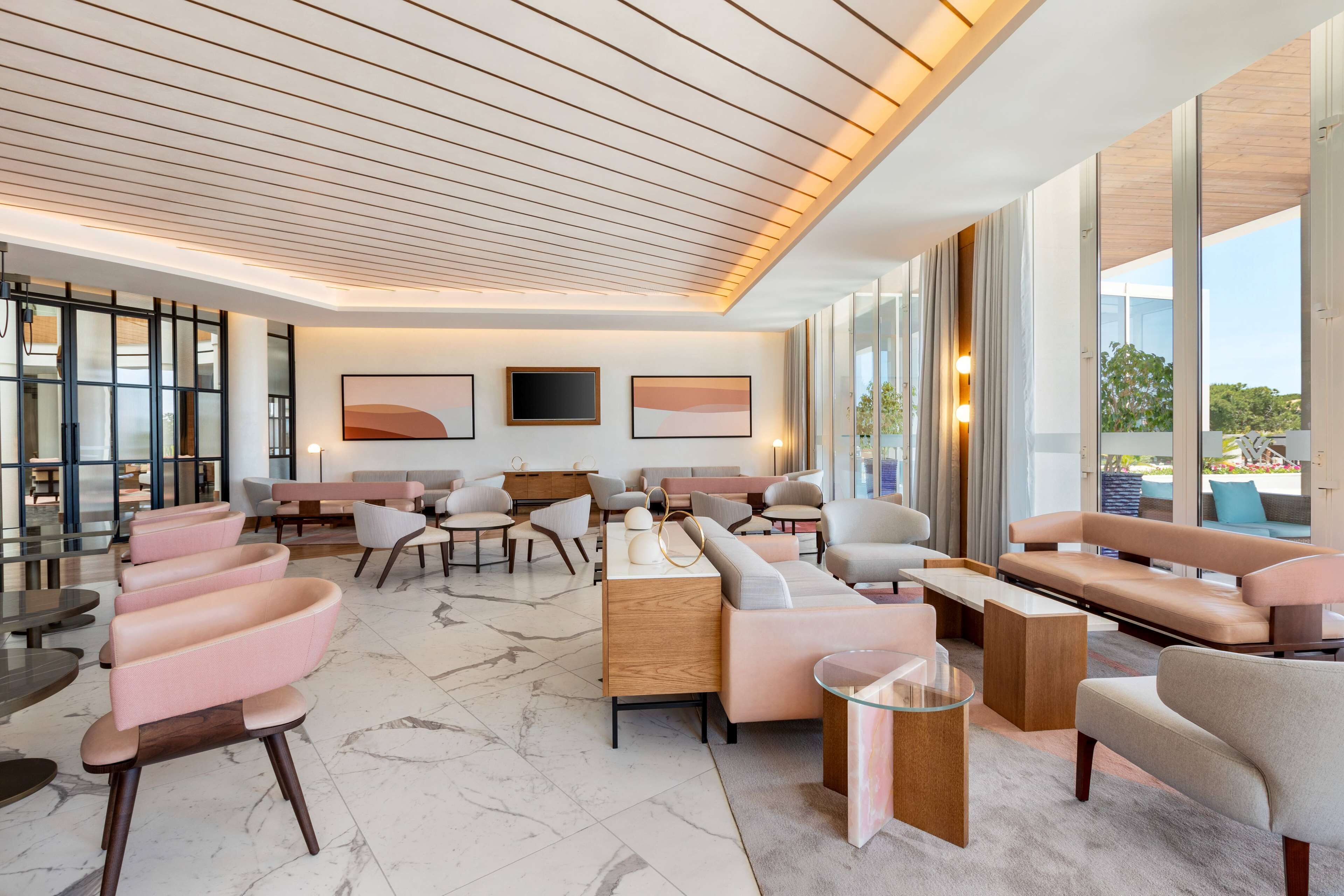 A modern lounge area with a mix of seating options, including sofas and chairs, arranged around a central coffee table. Large windows provide views of the outdoors.