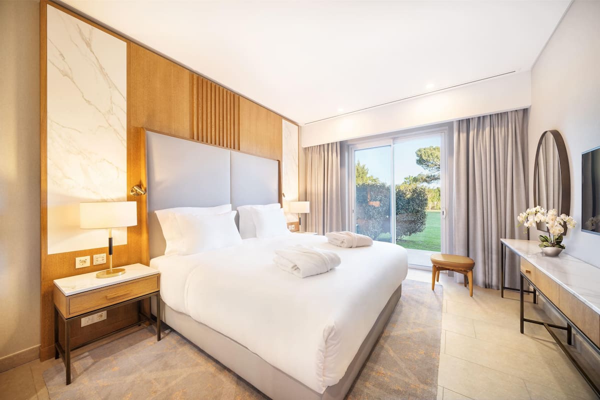 A modern hotel room with a large bed, a wooden headboard, a bedside table with a lamp, a small stool, a desk with a vase of flowers, and a large window with curtains.