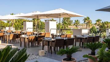 An outdoor dining area with wicker tables and chairs, large umbrellas, and potted plants.