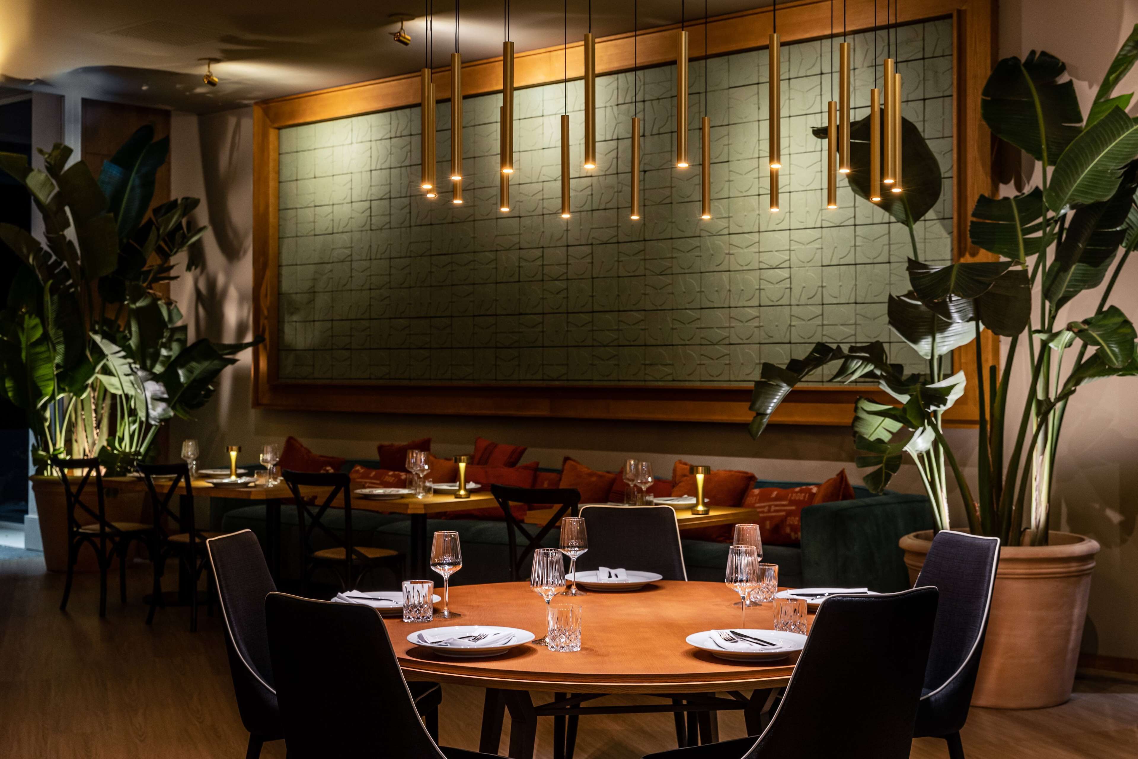 A restaurant with a round table set for dining, multiple chairs, hanging pendant lights, and large potted plants.