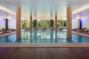 A modern indoor swimming pool with clear glass walls, wooden flooring, and multiple lounge areas with chairs. - Wyndham Grand Algarve Quinta do Lago (Loulé)
