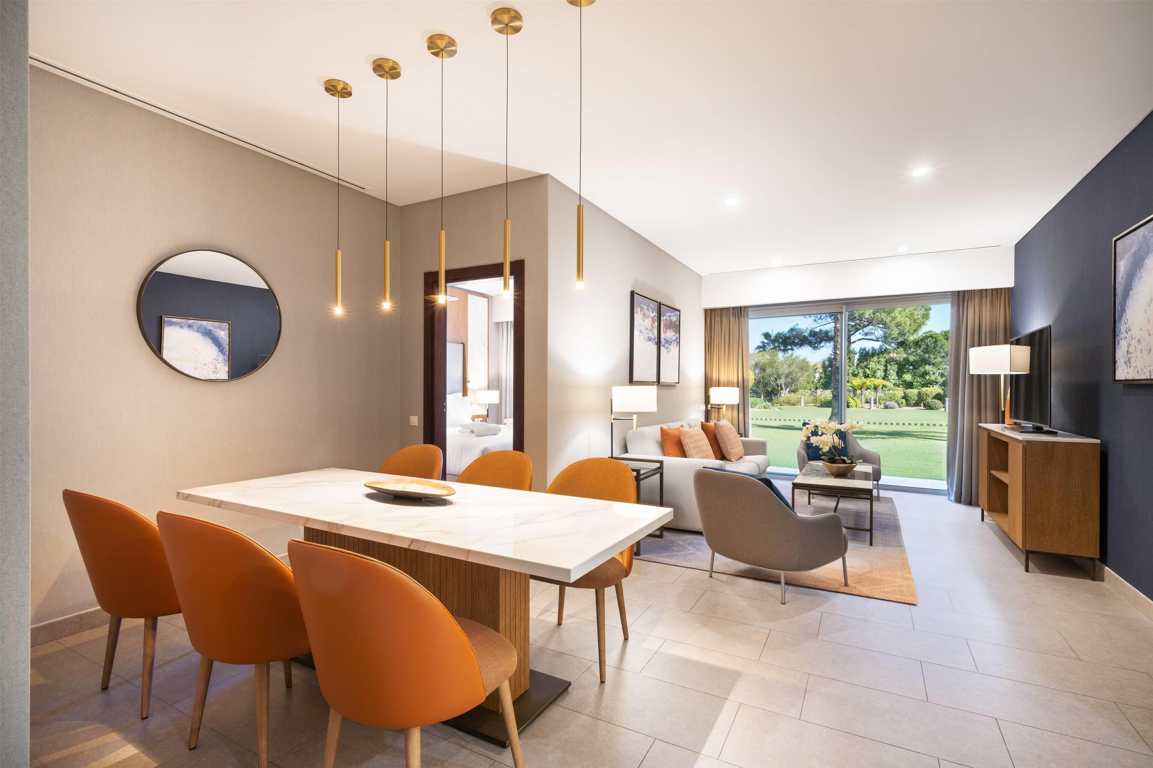 A modern dining area with a large table, orange chairs, and pendant lighting. There is a view of a garden through large windows.