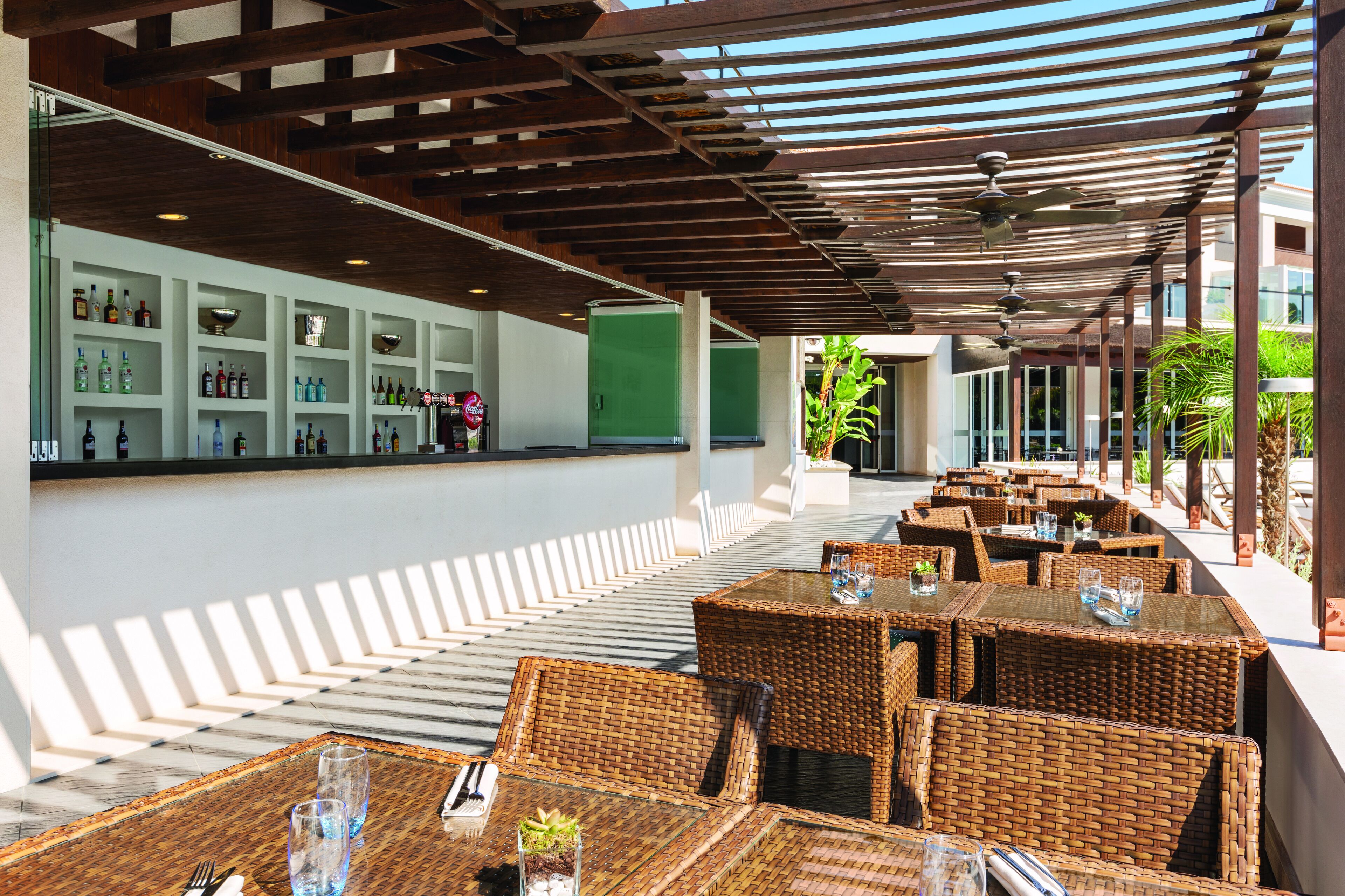 A covered outdoor seating area with wicker tables and chairs, a bar with shelves of bottles, and a green wall in the background.