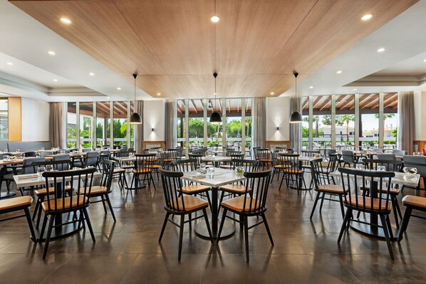 Un vaste espace dédié à la salle à manger, avec de nombreuses tables et chaises, de grandes fenêtres donnant sur un paysage verdoyant et un plafond en bois.