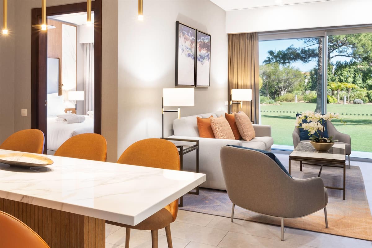 A modern hotel room with a dining area, a sofa with orange and beige cushions, a coffee table, and a view of a well-manicured garden.