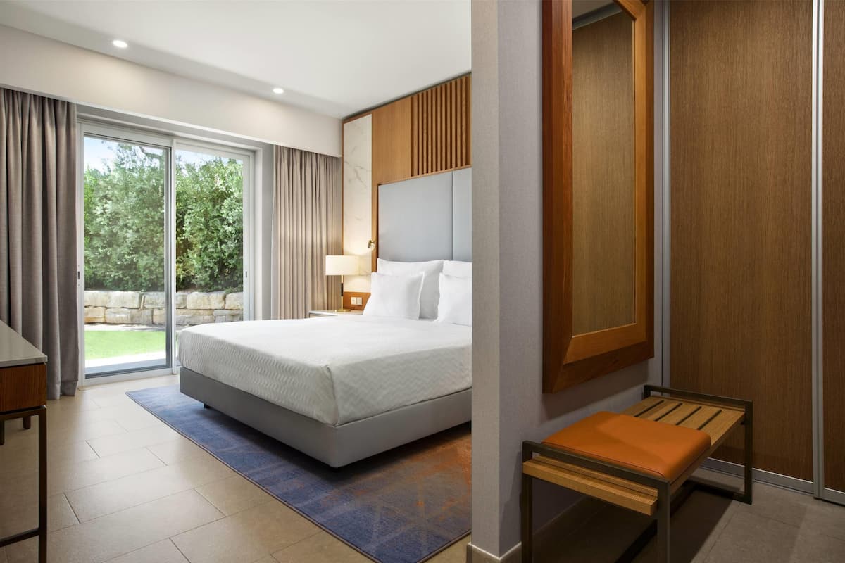 A modern hotel room with a large bed, a bench, a desk, and a view of the outdoors through a sliding door.
