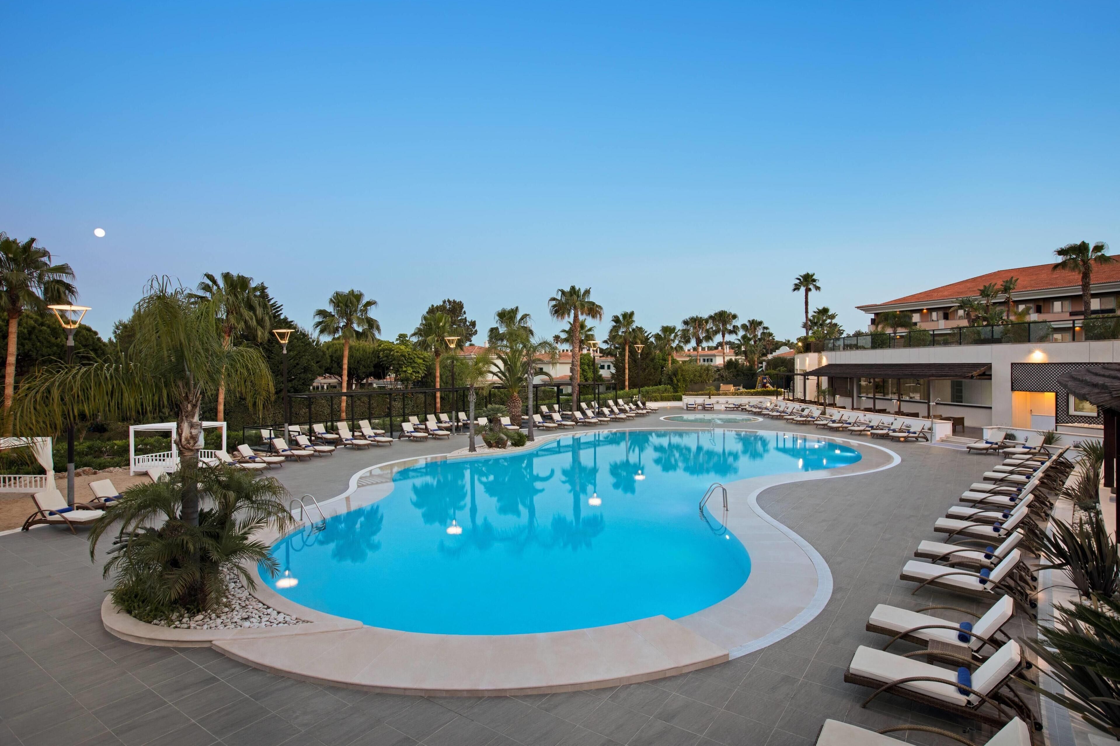 A large, curved swimming pool surrounded by lounge chairs and palm trees, with a building in the background.