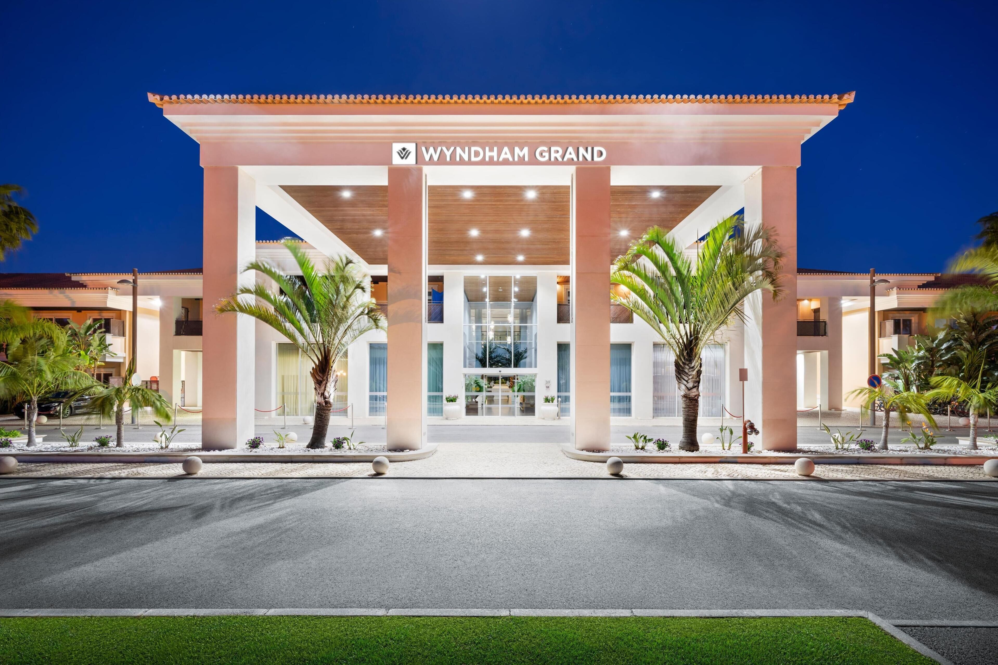 The Wyndham Grand hotel entrance with a modern facade, palm trees, and a well-lit driveway.
