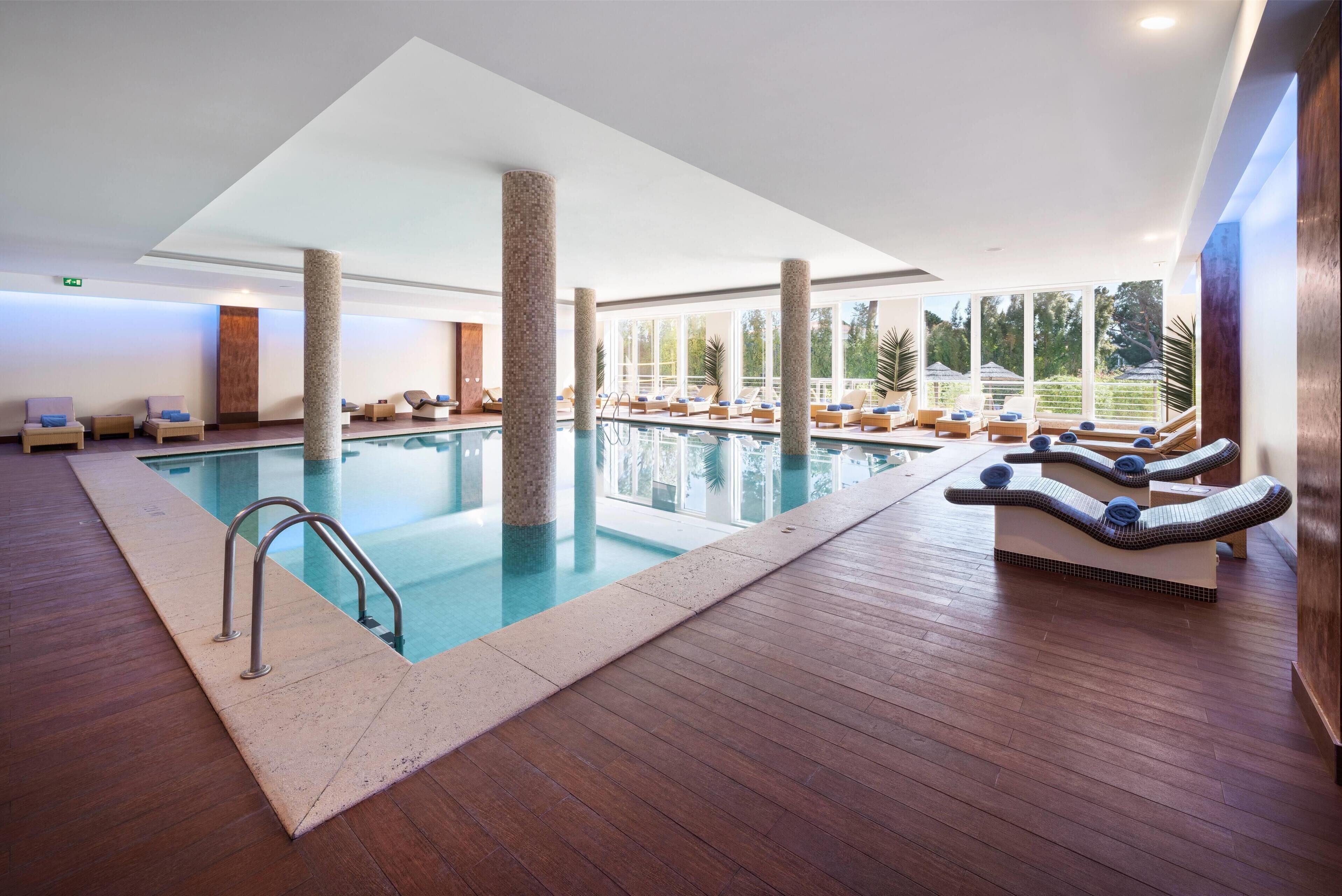 Una piscina interior moderna con suelo de madera, sillones de descanso y grandes ventanales con vistas al exterior.