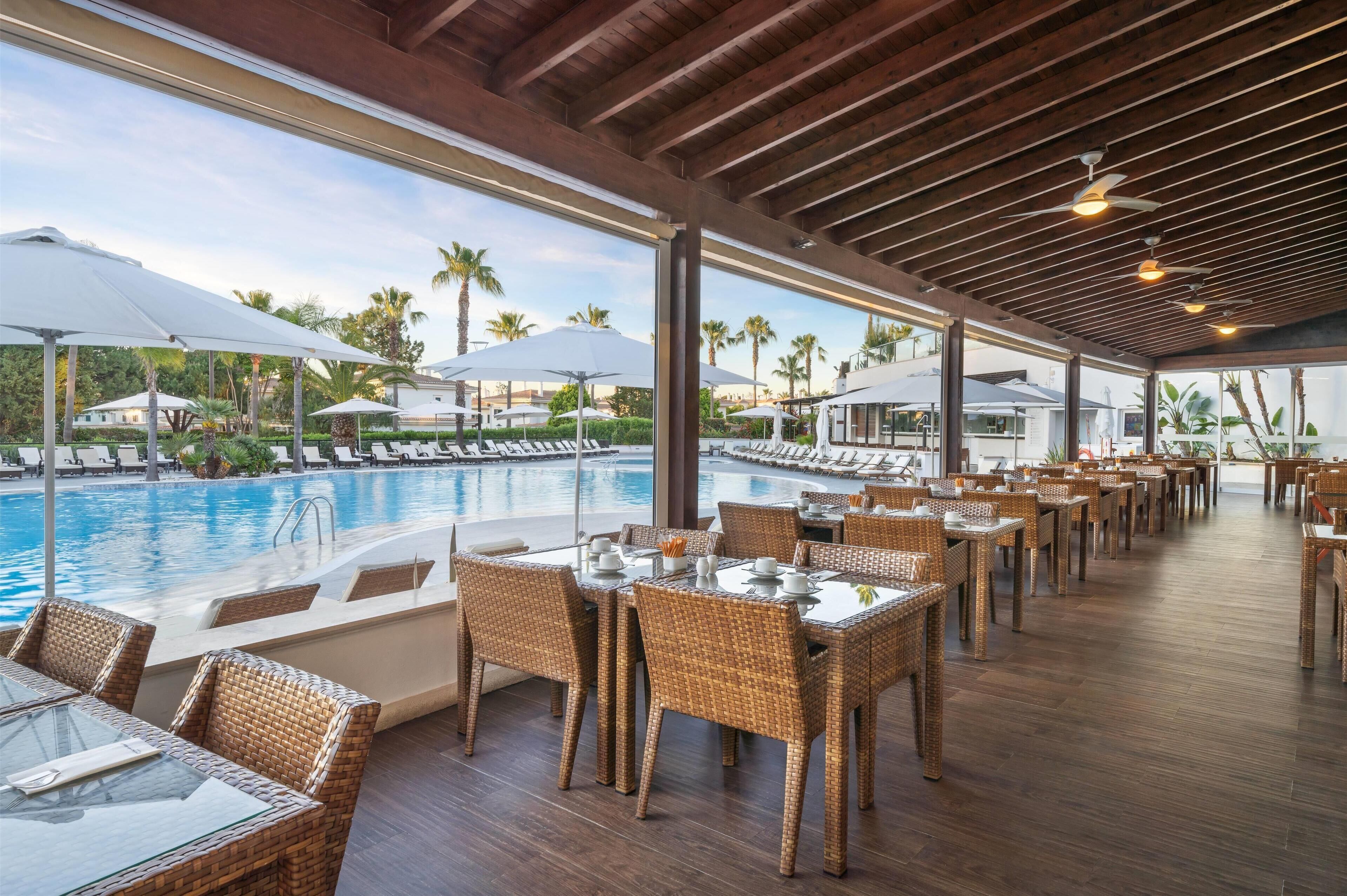 Ein Poolrestaurant mit Korbmöbeln, großen Sonnenschirmen und Blick auf den Pool und Palmen.